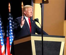 Donald Trump speaking at a lectern with US flags behind him.